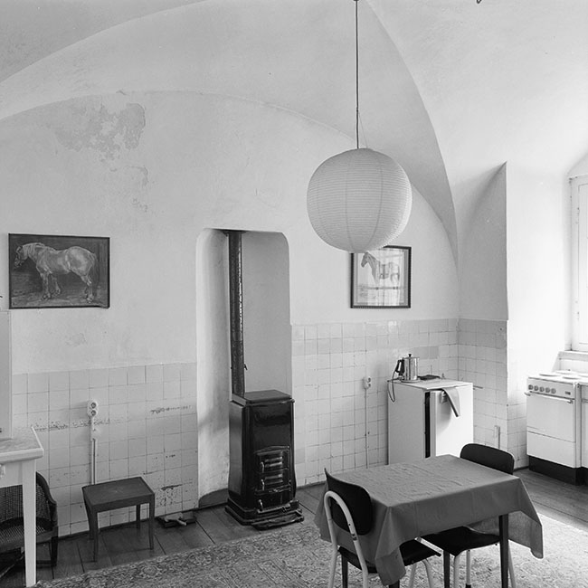 Zwartwitfoto van een ruimte met een gewelfd plafond. Tegen de onderste meter van de wanden zitten witte tegeltjes. Rechts staat een tafelmodelkoelkast en rechts voor het raam een oud fornuis. Midden in de ruimte hangt een ronde rijstpapieren lamp. Midden in de achterwand zit een nis met daarin een zwarte kachel. Tegen de muur hangt aan iedere kant van de nis een afbeelding van een paard. In het midden staat een kleine tafel met twee stoelen.