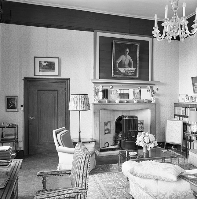 Zwartwitfoto van een zitkamer. Het licht komt van links, in de muur waar we tegenaan kijken zit links een deur en rechts een schouw met een ronde kachel. Boven de schouw hangt een portret van een 20ste eeuwse militair aan tafel met een boek voor zich. Boven de deur een afbeelding van Kasteel Amerongen vanuit het Zuiden en links naast de deur een gravure van een 17e eeuwse militair. Links voor de schouw een leunstoel met een schemerlamp, daarvoor een glazen koffietafel met een bosje bloemen. Rechts een lage boekenkast. Rechts op de voorgrond een bank en links een bureau. Rechts boven een glazen kroonluchter.