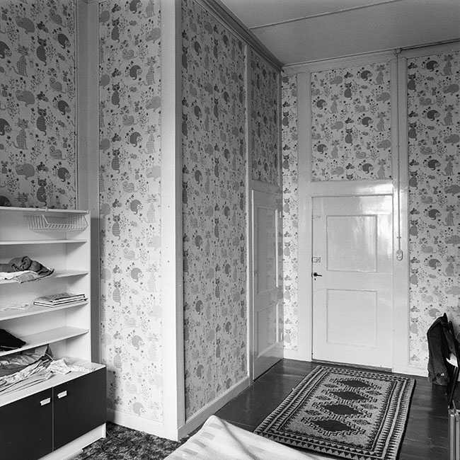 Zwartwitfoto van een kamer met een kattenbang. Links tegen de muur staat een kastje, rechts zien we nog net een stoel. Tegenover ons de deur en links daarvan een grote ingebouwde kast.