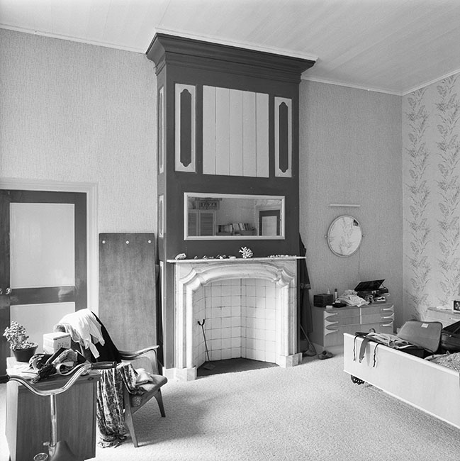 Zwartwitfoto van een kamer met een lege, marmeren schouw, de schoorsteenmantel is van hout met daarin een spiegel verwerkt. Rechts naast de schouw staat een laag kastje met een ronde spiegel erboven, rechts een soort gevederd behang met daarvoor nog een stukje van een tweepersoonsbed. Links naast de schouw een deur met daarvoor een stoel waarop kleren liggen, een klein vierkant kastje met daarop een plant en spulletkes. Op de voorgrond het stuur van een hometrainer.