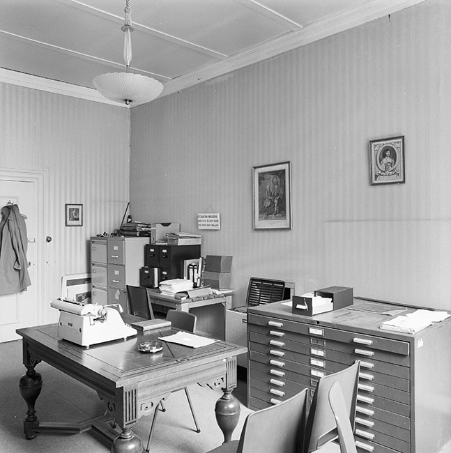 Zwartwitfoto van een kantoor met stevige eikenhouten tafel met daarop een grote typmachine een boek, een envelop en een asbak. De muren zijn behangen met een streepjesbehang en er zit een systeemplafond in. Twgen de achterwand staan archiefkasten in allerlei soorten en maten. Tegen de achterwand een deur met daaraan een kapstok met jassen. Aan de lange wand hangen twee prenten. Het behang heeft diverse verkleurde plekken.