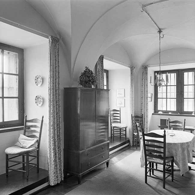 Zwartwitfoto van een ruimte met een gewelfd plafond en drie ramen in diepe nissen. In de nissen staan stoelen en voor de nissen hangt een gordijn. Tussen de nissen links staat een eenvoudige afgesloten kast. In het midden staat een tafel met afgeronde hoeken en twee stoelen. Aan het plafond hangt een eenvoudige luchter.