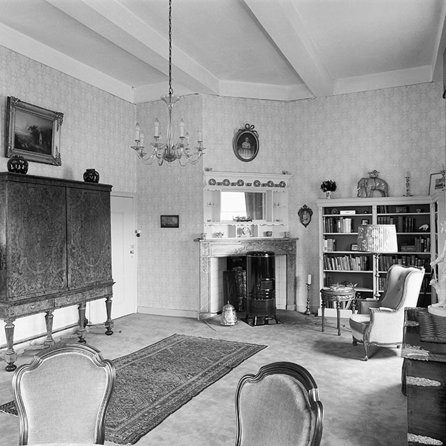 Een zwartwitfoto van een woonkamer. Het ligt komt van rechts, maar we zien geen ramen. Links staat tegen de muur een kabinet en rechtsdaarvan is een deur. Recht tegenover een schouw met daarin een hoge ronde kachel. Tegen de wand ernaast een boekenkast. Voor de boekenkast een leunstoel met schemerlamp en een klein rond bijzet tafeltje. Rechts zien we nog net de voorkant van een kist waarop we een deel van een beeldje zien. Op de voorgrond twee eetkamer stoelen. Op de vloer een tapijt.