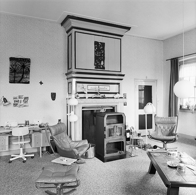 Zwartwit foto van een ruimte met een grote schouw waarvoor een stevige houtkachel staat. Voor de kachel staat een messing haardstel en twee leren leunstoelen, met ronde staande lampen. Links van de schouw staat een bureautje met daarboven wat foto's tegen de muur geplakt. Rechts van van de schouw een deur, rechts in de hoek een raam met plantjes op de vensterbank. Rechts voor een eiken koffietafel met een tegelblad. Op de tafel staan een koektrommel en wat snuisterijtjes. Bij de ene leunstoel ligt een breiwerk op de grond. De vloer is bedekt met gemeleerd tapijt.
