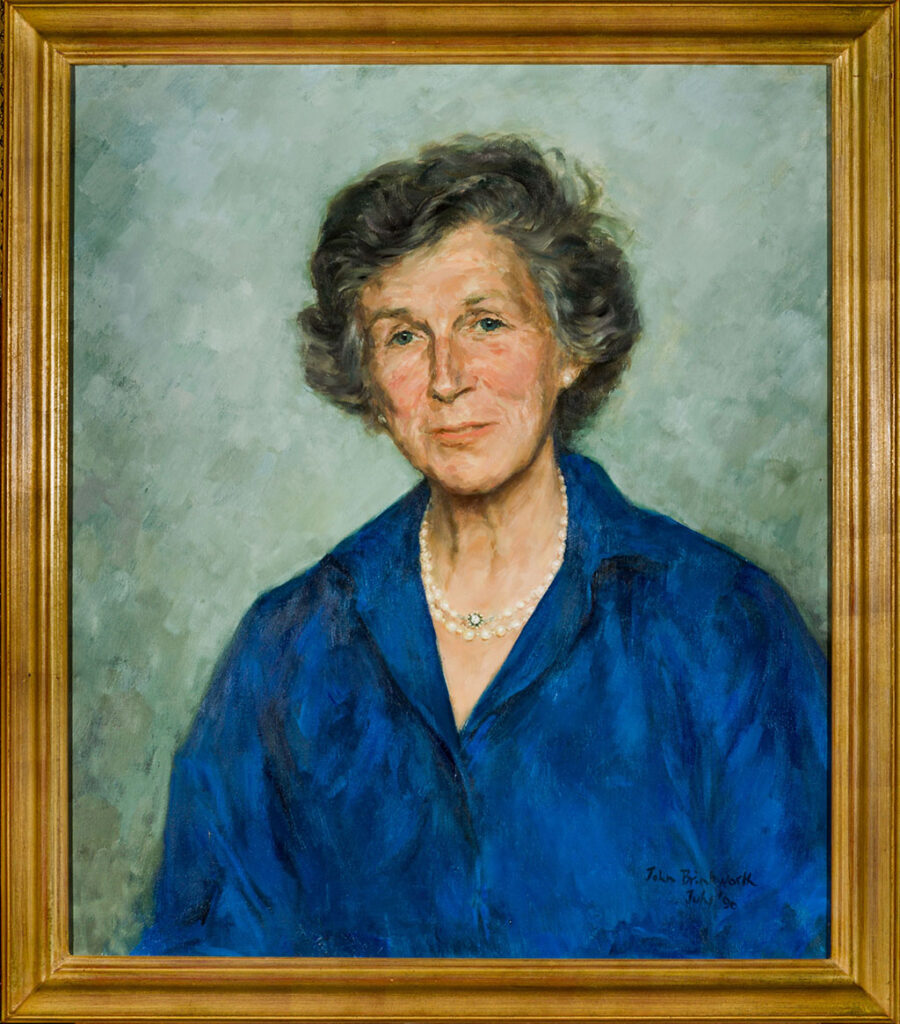 Een vriendelijk ogende dame met kort golvend donker haar met hier een daar een grijze lok. Ze draagt een blauwe blouse en een parelketting.