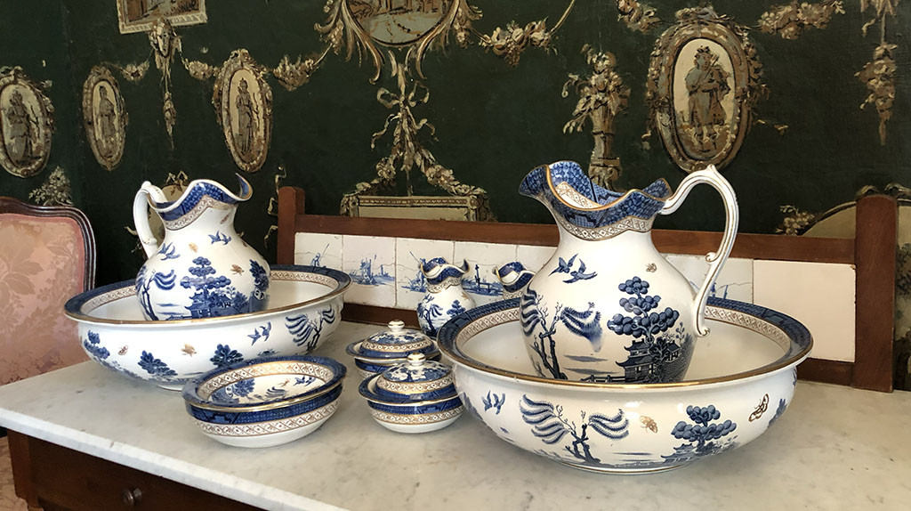 Voor een druk behang staat een tafel met een marmeren blad. Daarop een lampetset van wit aardewerk met een blauw landschapsmotief. Op tafel staan twee waskommen met in iedere komt een lampetkan, twee ronde zeepdoosjes, een rond schaaltje en verscholen achter de voorstel waskom staan twee vaasjes.