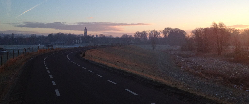 Een heiig landschap net voor zonsopgang, de bomen zijn kaal en op het weiland ligt rijp. Midden voor een asfalt weg op een dijk die een bocht naar rechts maakt. In de verte het dorp met in het midden de kerktoren. Aan de rechterkant zie je als je heel goed kijkt tussen de bomen de contouren van het kasteel.