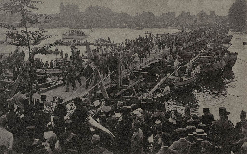 Zwartwit foto van de overtocht over de Schipbrug bij Dordrecht van het 1' Battillon, 15' Regiment Infanterie van het Korps Pontonniers tijdens de mobilisatie. Op de voorgrond speelt een militaire kapel. Op de achtergrond de stad.