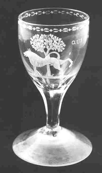Helder, kleurloos glas. Conisch voetje. Gefaceteerde stam, die overgaat in een tulpvormige cuppa, waarop gegraveerd een oranje boompje, waartegen een hond plast. Het opschrift "Honi soit qui mal y p..."