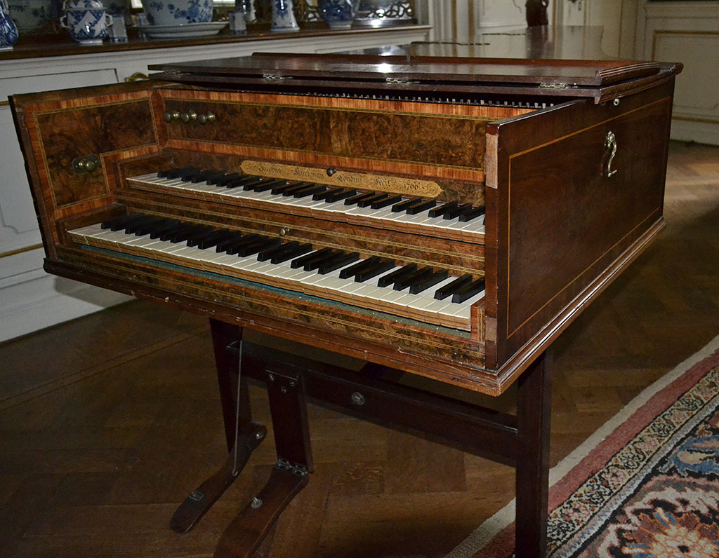 Een houten klavecimbel met twee klavieren. Links zitten drie knoppen om de aanslag en het volume te bepalen. Het instrument heeft een houten pedaal.