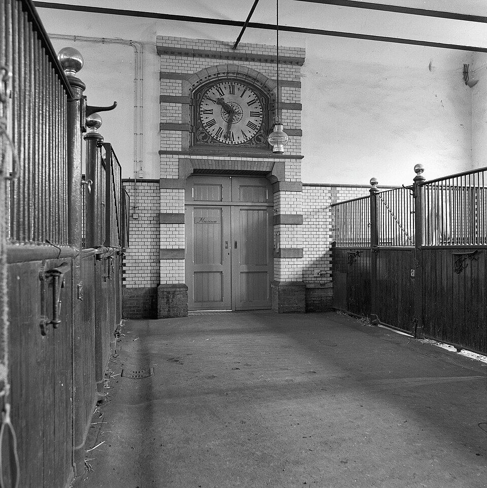 In een brede ruimte zitten aan weerszijden paardenboxen. Het onderste beschot is van hout, daarboven traliewerk. In de box rechts achter staat een schimmel. Boven de deur in het midden is een klok in de muur verwerkt. Op de deur staat in sierlijke letters "Museum".