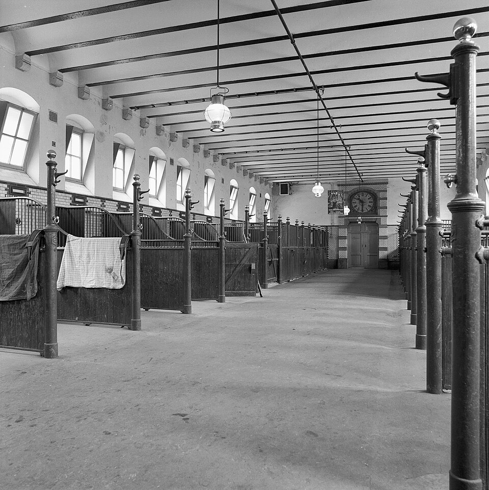 In een brede ruimte met hoge ramen zijn aan weerszijden stalruimtes afgebakend met schotten en tralies die vanaf de achterkant oplopen tot aan de muur. Op de achtergrond is een aantal boxen te zien en boven de deur in het midden is een hele grote klok verwerkt in de muur.
