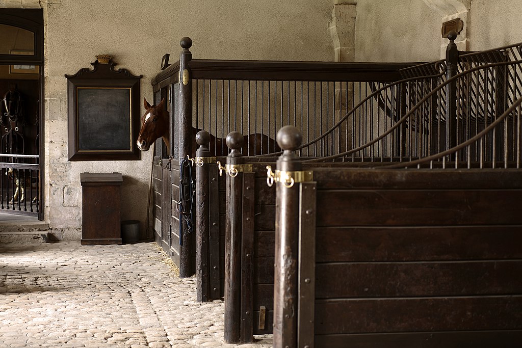 Een oude stal met rechts achter een afgesloten deel met hoge tralies en een open ruimte boven de deur, in deze box staat een paard. Hiervoor zijn drie stands met half hoge schotten en op de schotten tralies die hoger worden richting de muur. Achter tegen de muur een krijtbord met daaronder een kist, op de vloer liggen kinderkopjes.