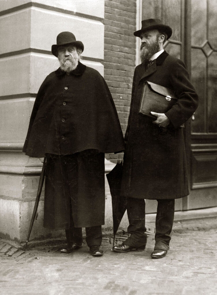Zwartwit foto van Twee mannen, links duidelijk de oudere met een lange zwarte jas en daaroverheen een zwarte cape. In zijn rechterhand een strak opgerolde paraplu. Hij heeft een grijze volle baard en snor en hij draagt een bolhoed. Links een jongere en iets grotere man. Hij draagt een lange zwarte jas en een zwarte broek. In zijn rechterhand een gesloten paraplu, onder zijn linkerarm draagt hij een leren tas. Ook hij heeft een baard en snor, maar donker en rond de mond iets grijzend. Op zijn hoofd een zwarte gleufhoed.