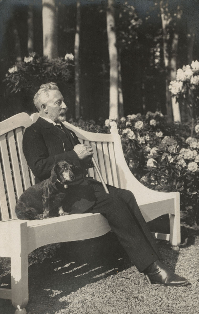 Op een wit bankje in een tuin zit een oudere man met kort grijs haar, een snor en een sikje. Hij draagt een zwart pak en heeft een stok in zijn handen die tussen zijn benen staat. Naast hem op het bankje zit een tekkel.