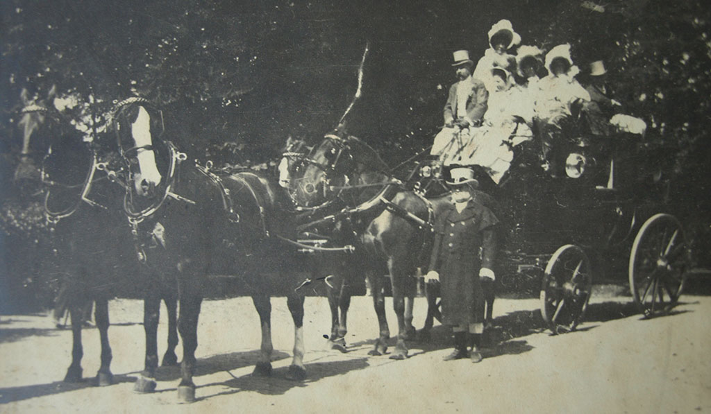 Zwartwitfoto van een koets met ervoor vier paarden. Op de koets zit een man in een grijs pak met een hoge hoed die de leidsels vast houdt, naast hem zit een vrouw in wit met een hoedje op. Bovenop de koets zitten nog drie vrouwen in wit met hoedjes en achterop een man met een hoge hoed. Naast de paarden staat een man in een lange donkere jas en laarzen met een hoge hoed op.