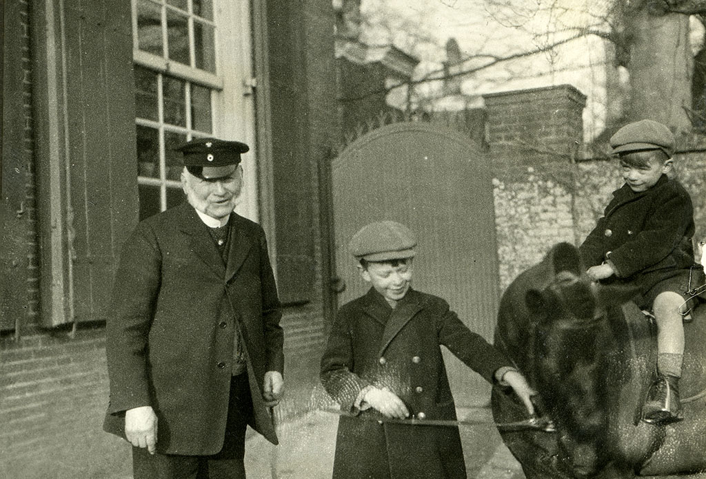 Voor een poort in de muur staat een oudere man met twee jongens en een pony. De oudere man is in het zwart, heeft een pet op en kijkt naar de camera. Naast hem staat een jongen van een jaar of acht à negen die een zwarte pony bij de teugels pakt. Op de pony zit een kleinere jongen van een jaar of zes.