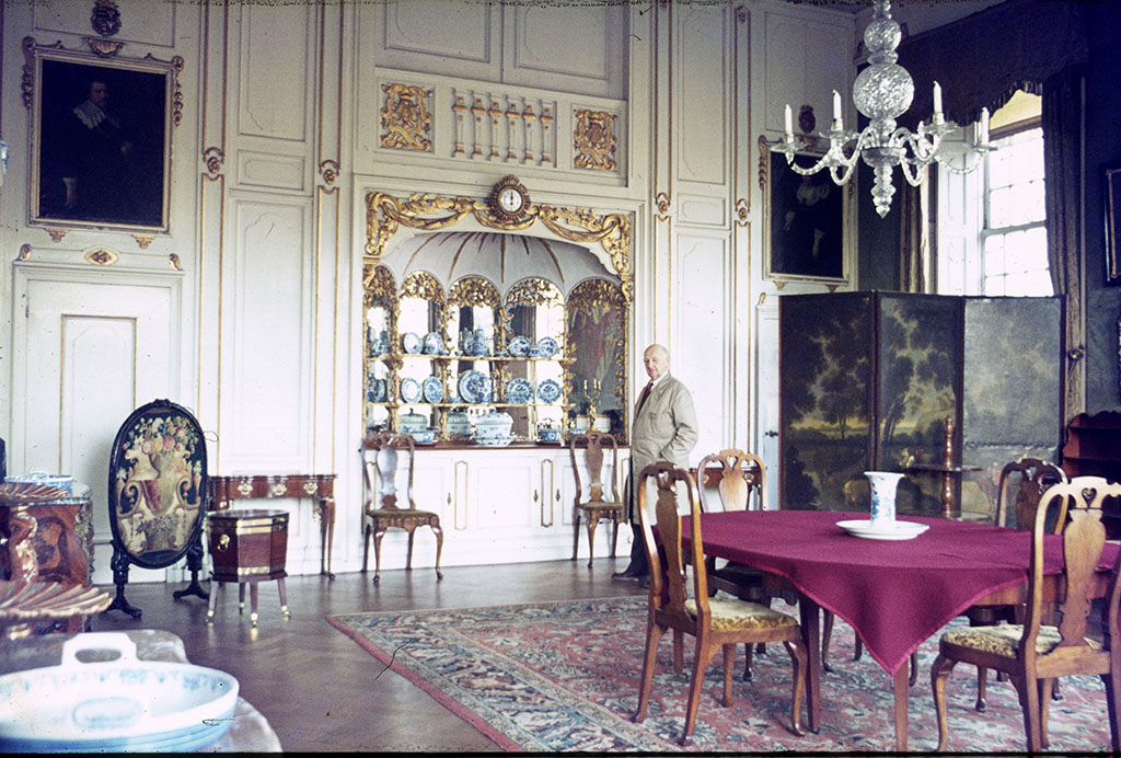 Kleurenfoto van de eetkamer. In het midden een ovalen eettafel met vier stoelen op een tapijt, erboven een kroonluchter met kaarsen. De achterwand is gelambrizeerd met daarin een buffet met Chinees porselein. In de wand zitten twee deuren, voor de deur rechts achterin staat een kamerscherm. Voor het buffet staat een oudere man in een stofjas.