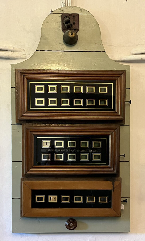 Een samengesteld bellenbord. Boven een oude elektrische bel, daaronder drie houten kasten met verschillende hoeveelheden vakjes. Onder de houten borden een ronde houten knop.