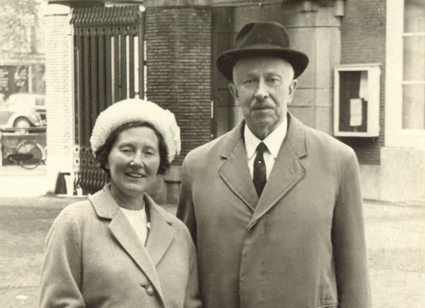 Zwartwit foto van een een oudere heer (rechts) met een hoed op en een jas met daaronder een overhemd en stropdas. Links van hem een vrouw met donker haar en een witte hoed. De vrouw lacht, de man kijkt serieus. Op de achtergrond een gebouw in een stad.