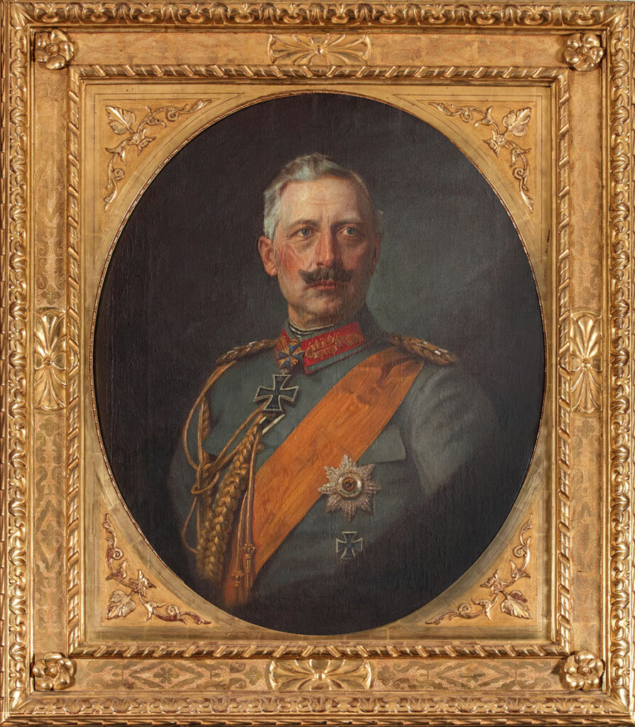 Ovaal busteportret van Keizer Wilhelm II tegen een donkere achtergrond in een zware vergulde lijst. De keizer zit met zijn lichaam naar links en kijkt weg naar rechts. Zijn grijze haar is naar achteren gekamd en de punten van zijn snor staan omhoog. Hij draagt een grijs uniform met een orde kraag met een oranje sjerp. Op zijn borst heeft hij de Orde van de Zwarte Adelaar en hij draagt het IJzeren Kruis.