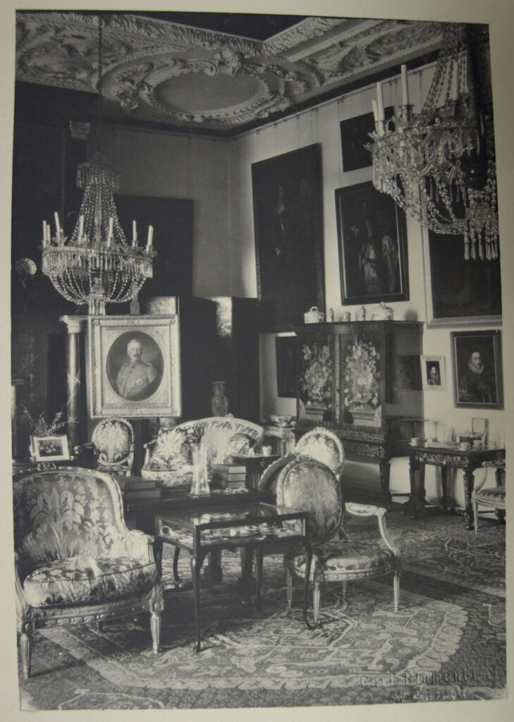 Zwartwitfoto met het interieur van de noordoosthoek van de grote zaal van Kasteel Amerongen: elegante stoelen en banken, bewerkte kabinetten, een versierd stucplafond, schilderijen, snuisterijen en kroonluchters. Bij de schouw staat een busteportret van Keizer Wilhelm II op een ezel.