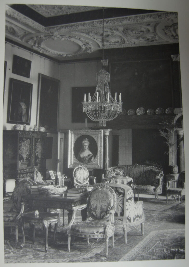 Zwartwitfoto met het interieur van de zuidoosthoek van de grote zaal van Kasteel Amerongen: elegante stoelen en banken, bewerkte kabinetten, een versierd stucplafond, schilderijen, boeken, snuisterijen en kroonluchters. Bij de schouw staat een busteportret van Keizerin Auguste Victoria op een ezel.