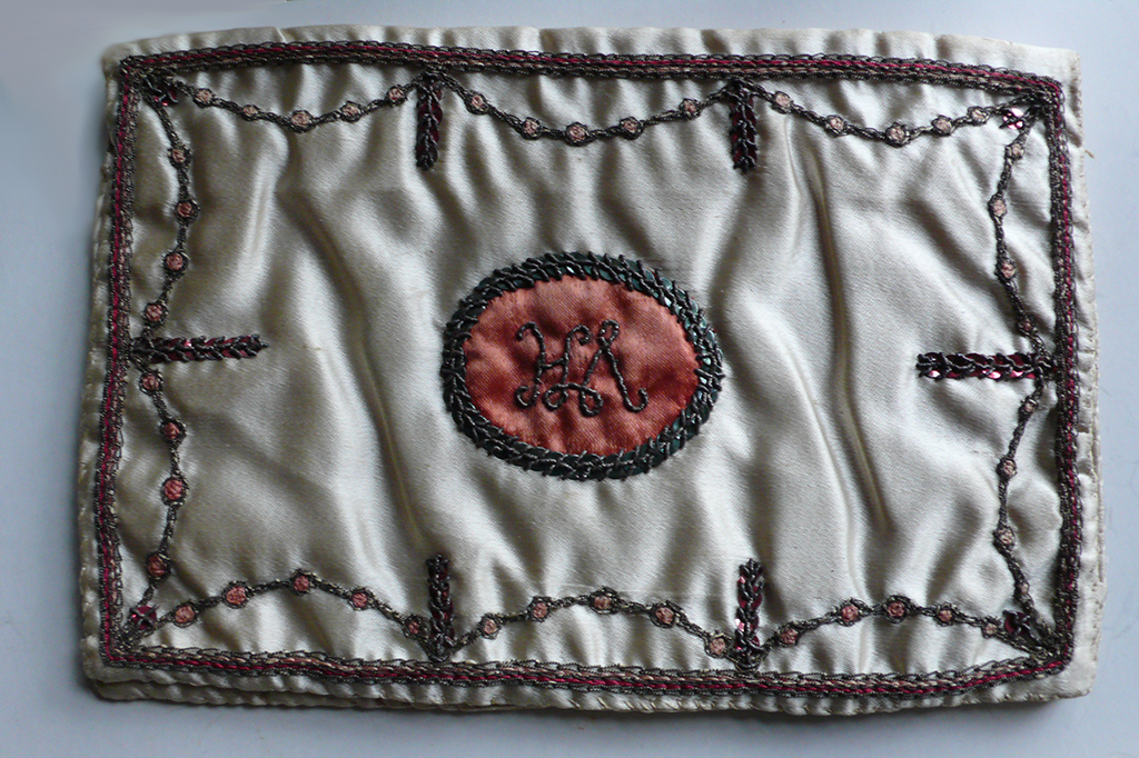 Een eenvoudig wit zijden etuitje geborduurd met een witte rand met guirlandes en gestoenen. In het midden een ovaal met een nog niet opgelost monogram.