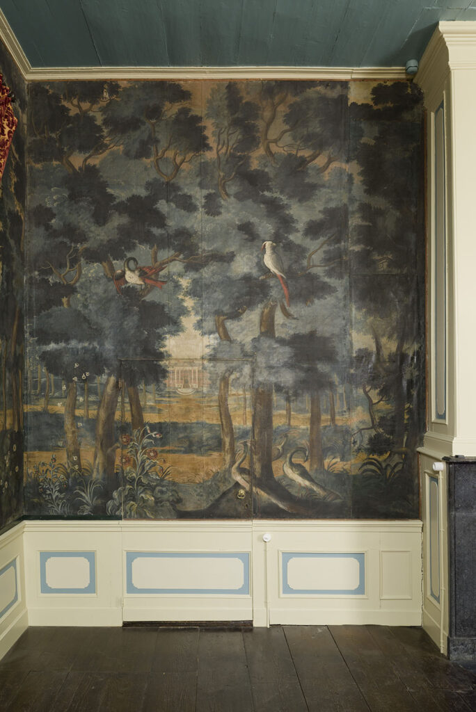 Een wand met een lage lambrisering met beige en lichtblauw, rechts zie je nog net de schouw. In het midden zit een deur, maar de beschildering loopt door over de deur. Aan de bovenste rand van de deur paleis soestdijk, op de voorgrond twee pauwen en een papegaai en een andere vogel in de bomen.