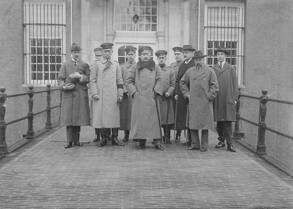 Zwartwitfoto van een groep mannen op de brug voor kasteel Amerongen. Alle mannen dragen lange jassen, de meesten militaire petten, de anderen een hoed. Ze hebben bijna allemaal snorren. De man links heeft een tekkel in zijn armen.