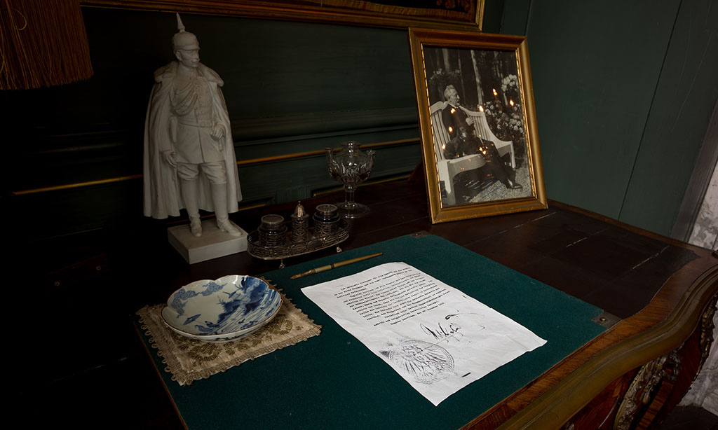 Foto van het blad van een bureau, een groen vilten schrijfblad met daarop een wit A4-tje met getypte tekst en een handtekening. Links ervan een geborduurd doekje en daarop een chinees schaaltje. Boven de het document ligt een pen. Op tafel staan verder een wit beeldje van Keizer Wilhelm II in militair uniform met Pickelhaube, een zilveren inktstel, een glas en een ingelijste foto van de keizer als oude man op een wit bankje met naast hem zijn tekkel.