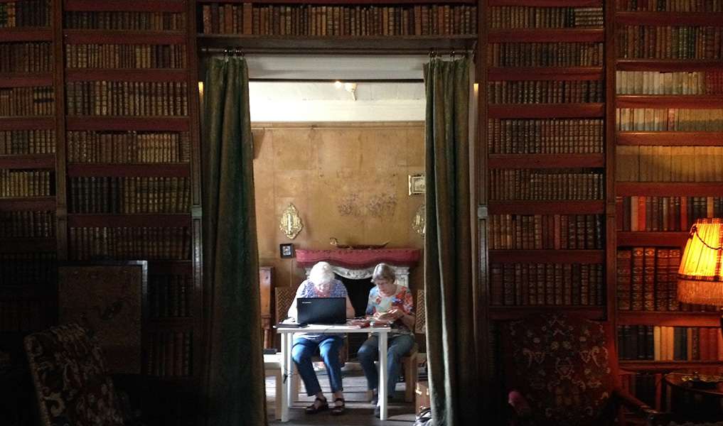 Tussen donkere boekenkasten zit een doorgang met twee gordijnen ervoor die open zijn. De kleine ruimte erachter is helder verlicht door daglicht van links. Achter een modern tafeltje zitten twee dames achter een laptop te werken.