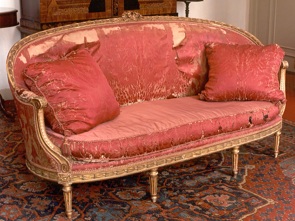 Een Lodewijk XV canapé: een bankje met een rondlopende hoge, rechte rugleuning/armleuningen. Het frame is van versierd verguld hout, de bekleding is van een sterk verouderd roze zijde-brokaat. Op de rugleuning is de stof voor een deel helemaal weg, voor het grootste deel zijn de patronen op de stof ook weggesleten, waar het zitkussen begint zie je nog een heel klein beetje van de helderheid van de oorspronkelijke kleur van het patroon. Het bankje heeft aan de voorkant vier dunne pootjes. Op de bank liggen twee losse kussens met de zelfde (versleten stof). Op de zitting zit een effen roze stof.