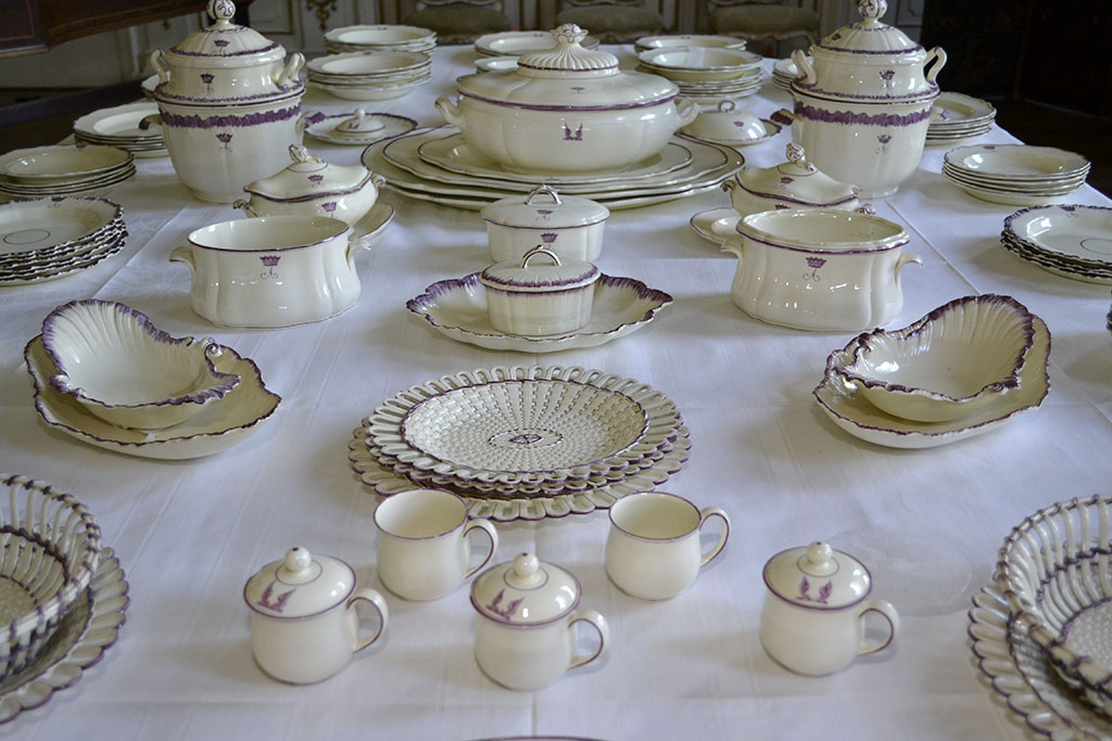 Op een tafel gedekt met een wit tafelkleed staan diverse onderdelen van een cremekleurig servies met paarse accenten. Op de voorgrond kopjes, bakjes, schaaltje met deksel, zonder deksel, platte schalen, borden…