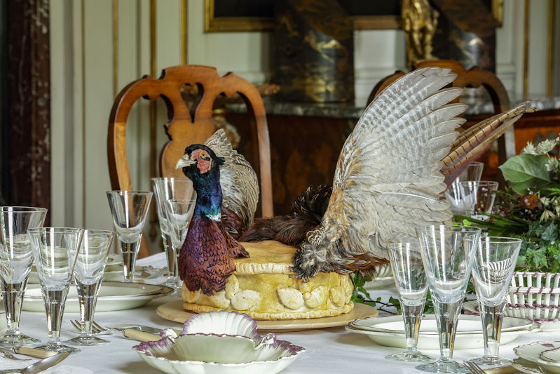 Op een tafel met glazen, servies en zilver staat een pastei waarop de kop van een opgezette fazant staat, zijn vleugels en zijn staart.