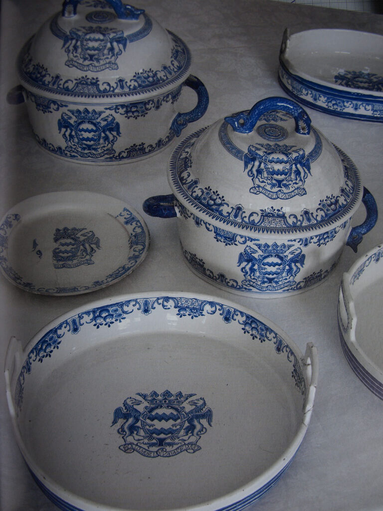 Onderdelen van een een wit-blauw servies op een tafel gedekt met damast. Er staan twee dekschalen met blauwe oren en een deksel, een grote ronde bak met handvatten (en twee deels zichtbaar) en een bordje gerestaureerd uit scherven.