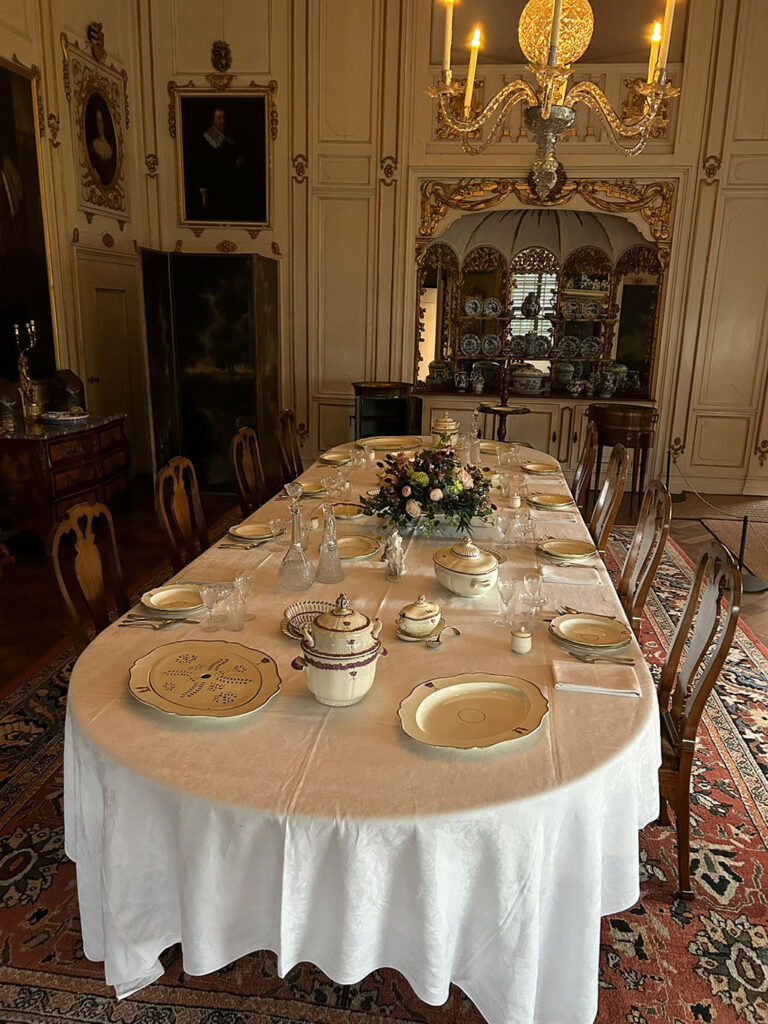 In de eetkamer staat een lange tafel met een wit tafelkleed een cremekleurig servies, glazen en een bloemstuk.