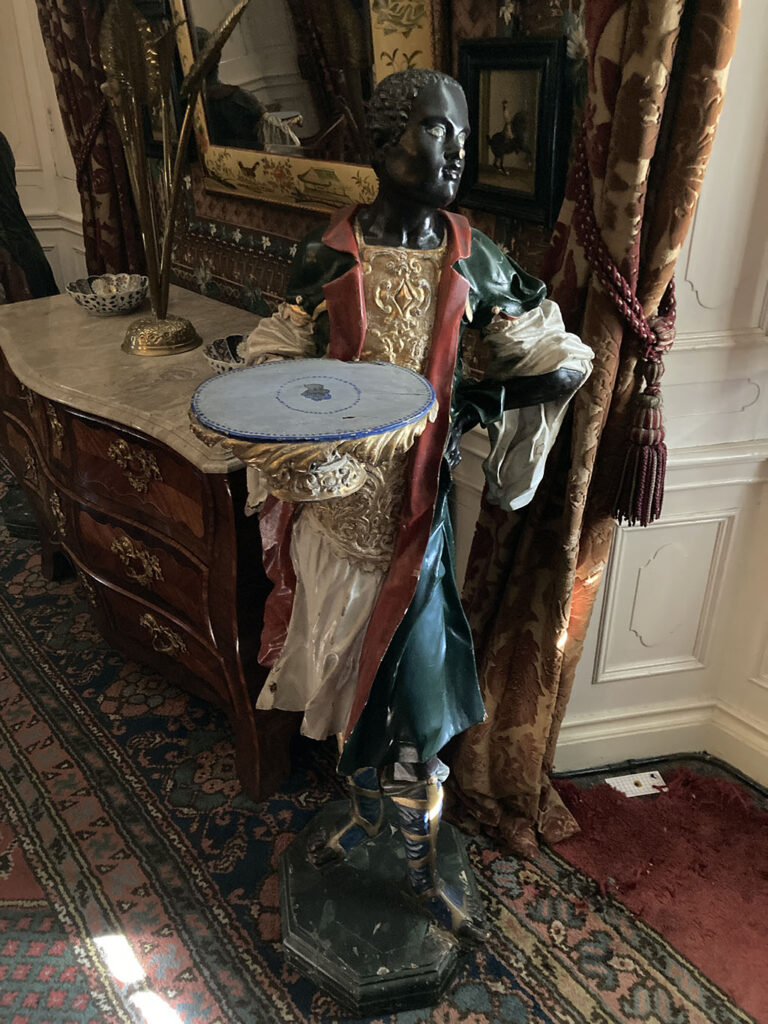 Een standbeeld van een zwarte jongen in elegante, ruimvallende kleding. Zijn linkerhand heeft hij in zijn zij, zijn rechterhand houdt een dienblad omhoog dat als bijzettafeltje gebruikt kan worden.
