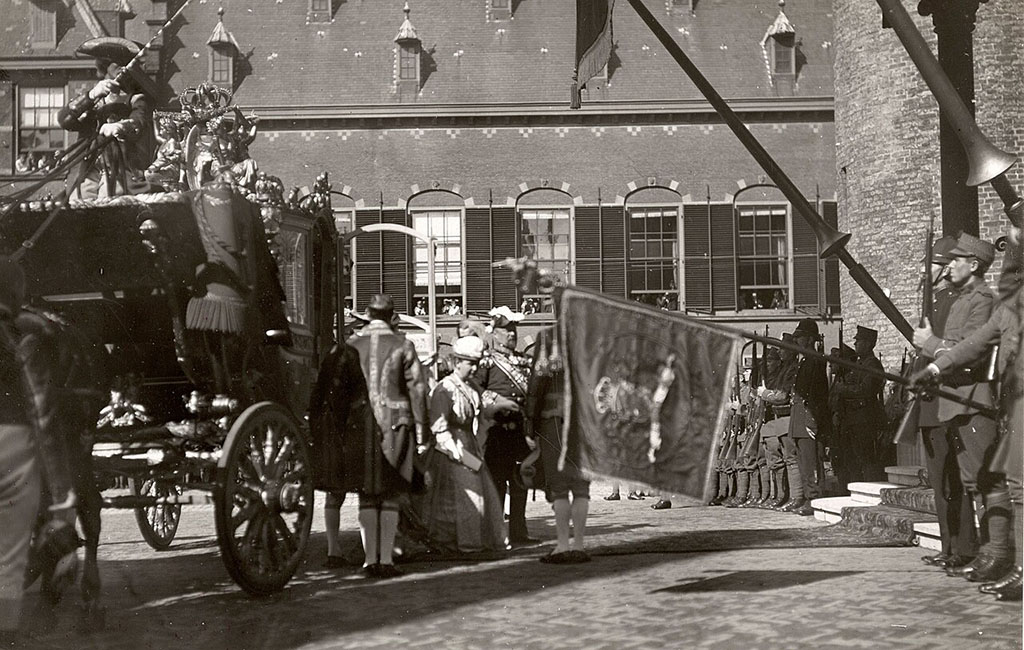 Zwartwit foto van links de gouden koets waar Koningin Wilhelmina met prins Hendrik uitstapt. Rechts de trap naar de ridderzaal. Daarvoor soldaten met lansen en één met een standaard. Op de achtergrond meer soldaten. Er hangen mensen uit de ramen op de achtergrond.