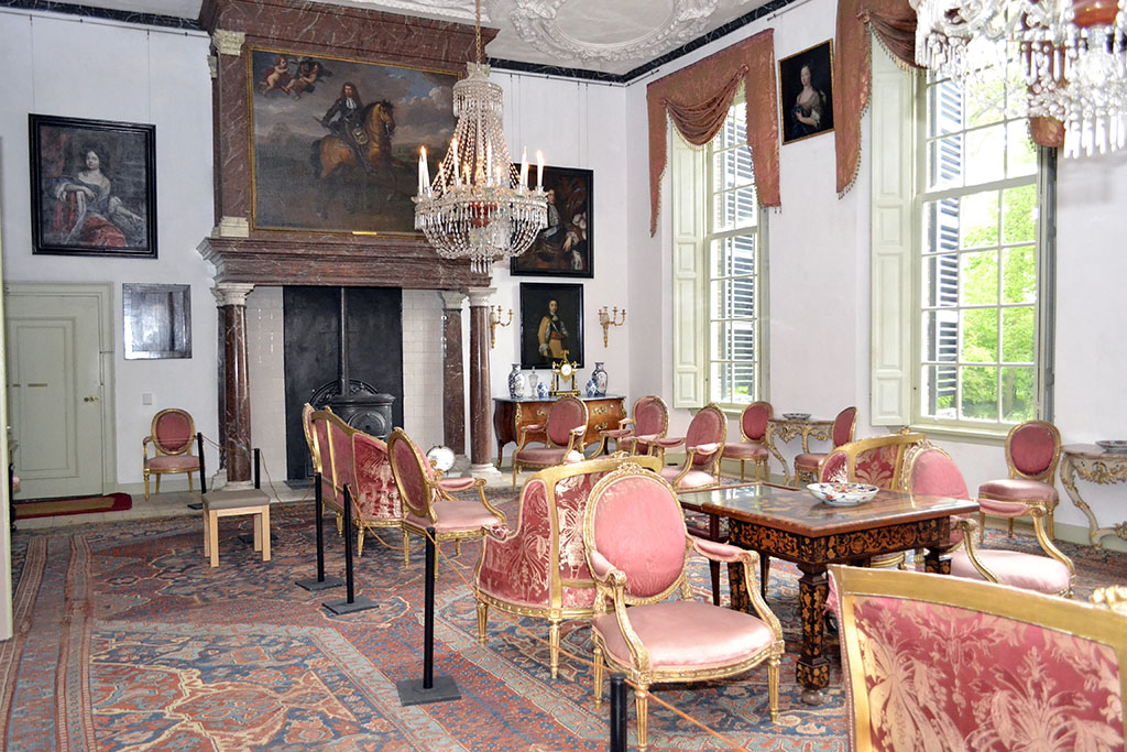 Grote zaal van Kasteel Amerongen. Een grote ruimte met hoge ramen. Aan de achterwand een roodbruin marmeren schouw met een portret van de Keurvorst van Brandenburg te paard. De wanden zijn wit en er hangen schilderijen. Linksachter een deur die dicht zit. Recht zijn zowel de binnen- als buitenluiken open, voor de ramen hangen alleen lambrequins aan de bovenkant van het raam. Het meubilair bestaat vooral uit losse stoelen en een enkele tafel en bank. Het meubilair staat vooral aan de rechterzijde van de kamer. Op ongeveer eenderde van de ruimte staan paaltjes met een koordje. Aan de lege zijde van het koord staat een lichthoutenkrukje met een kunstleren zitting.
