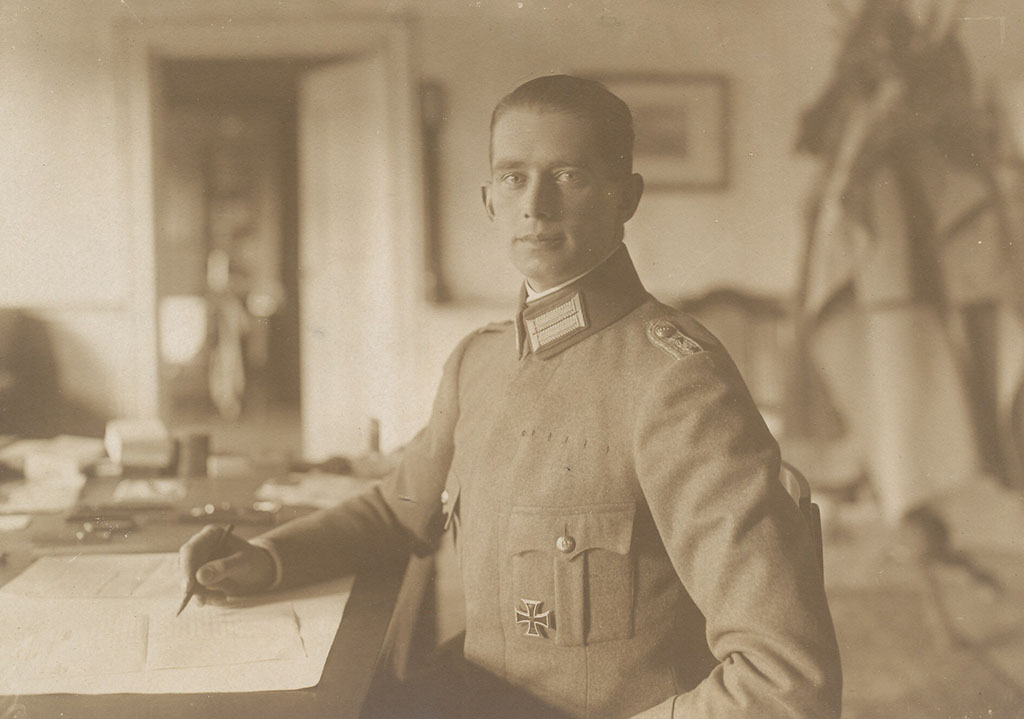Zwartwitfoto van een jonge man in een militair uniform aan een tafel waarop papieren liggen. In zijn rechterhand heeft hij een potlod. Op zijn linker borst draagt hij een Duits kruis.