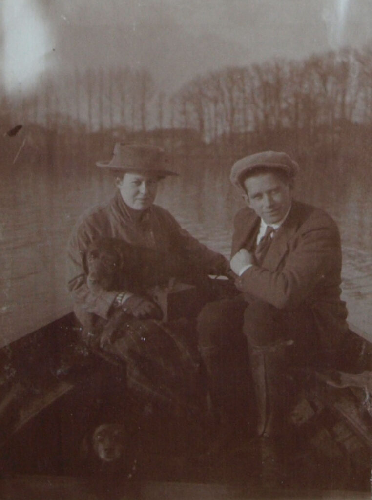 Zwartwitfoto van een man (Sigurd von Ilsemann) en een vrouw (Elisabeth van Aldenburg Bentinck) in een bootje. Ze varen op een groot water, op de achtergrond een rij hoge, kale bomen. Ze zijn beide warm aangekleed, zij draagt een hoed, hij een pet. Elisabeth heeft een hond op schoot en op de vorgrond zie je nog net een hondje dat in de camera kijkt.