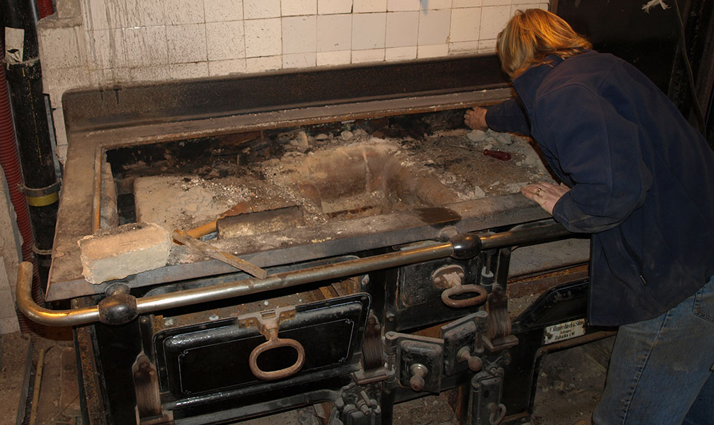 Een man staat voorover gebogen over een groot gietijzeren fornuis. De bovenkant is eraf en we zien het binnenwerk met gemetselde stenen.