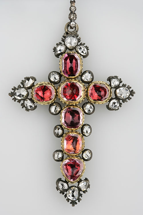 Een kruis bestaand uit zeven rood fonkelende robijnen, omgeven door twintig diamanten. Het geheel is gevat in goud en zilver.