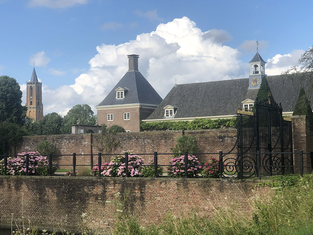 Een foto vanuit de tuin van kasteel Amerongen. Horizontaal loopt een kadewand met langs het pad daarboven roze hortensia's. Aan de andere kant van het pad zit een hoge muur. Rechts is het afgesloten met een zwart hek. Boven de muur uit zien we het dak van de stallen. Rechts de klokkenstoel, in het midden het aansluitende paviljoen. Helemaal links op de foto tussen de bomen de kerktoren van de Andrieskerk.