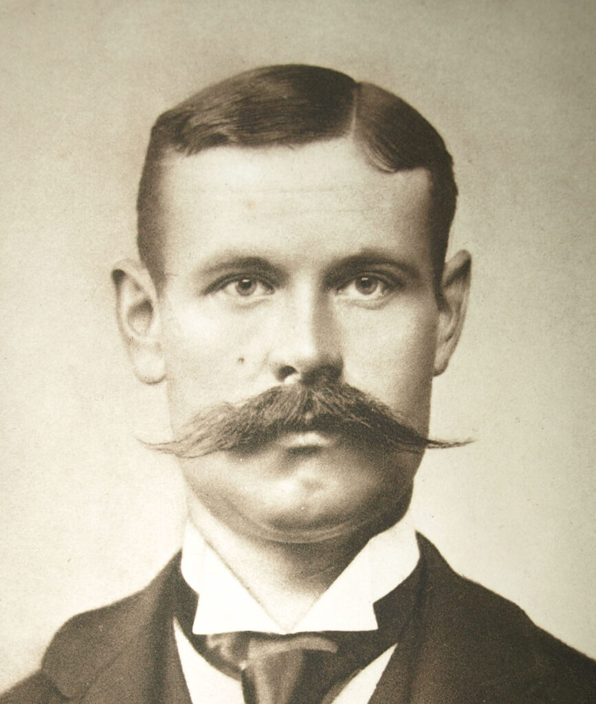 Portret en face van een jonge man met een kort kapsel en een brede snor. Hij draagt een pak.