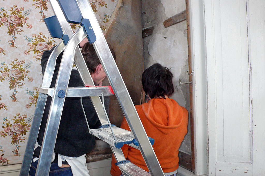 Een ladder staat voor twee mensen die in een hoek van de kamer kijken naar de muur die achter het behang zit dat opzij gehouden wordt. Op de muur zijn de latjes van de betengeling duidelijk te zien.