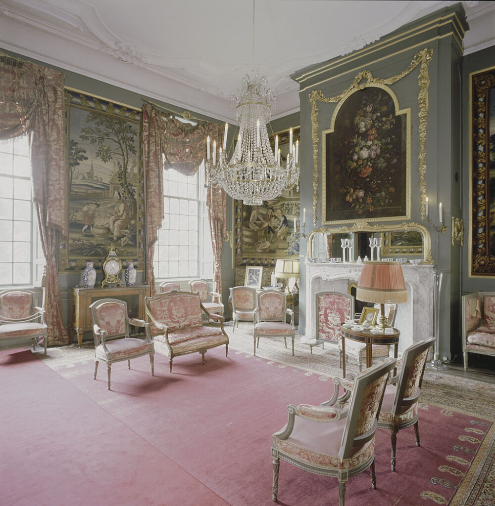 Een zonnige kamer met wandtapijten, groene lambrisering en een bloemstuk boven de schouw. Voor de ramen hangen gordijnen waar de gaten in gevallen zijn. Op de grond ligt een rood tapijt en er staan stoelen en een bankje met roodbrokaten bekleding.