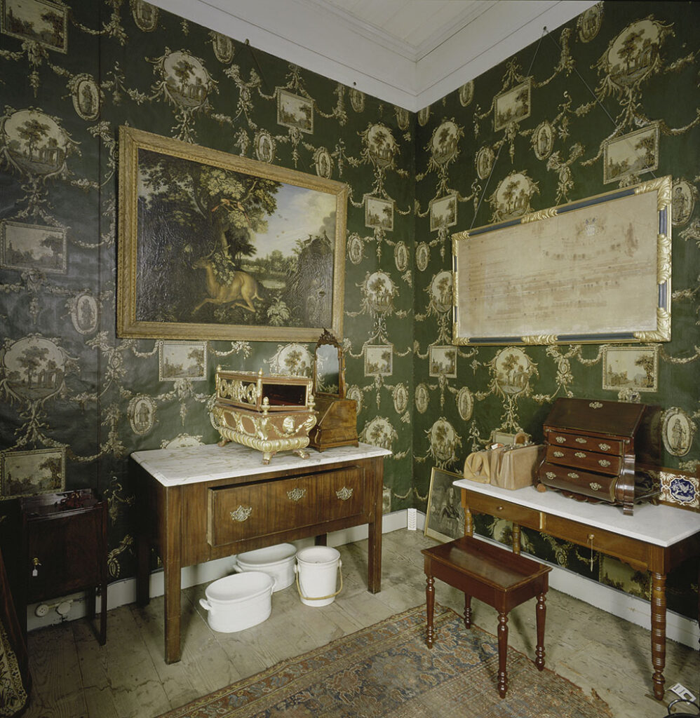 Foto van de hoek van een kamer. Op de muur een groen behang met daarop allemaal gedrukte medaillons en ingelijste prenten. Aan de linkerwand hangt een schilderij met een hert, aan de rechterkant een ingelijste stamboom. Onder elk schilderij staat een tafel met daarop kisten en kastjes. Bij de rechter tafel staat een kruk, onder de linker tafel staan keramieken bakken en een keramieken emmer. De vloer is van hout en we zien nog net een hoek van een versleten vloerkleed.