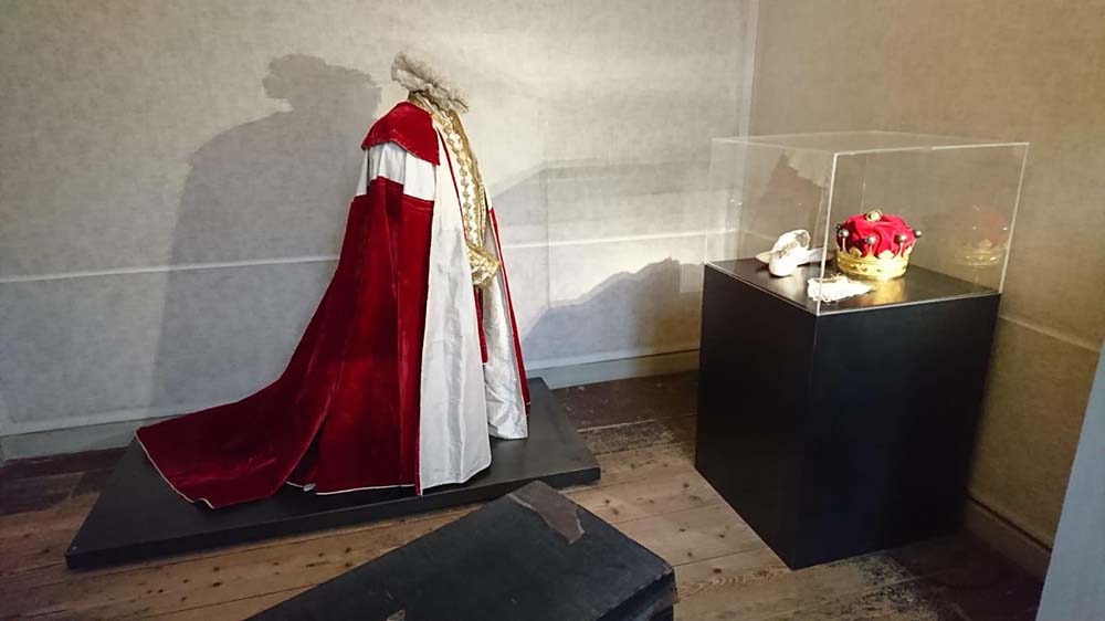 In een witte ruimte staat een paspop met een rode lange cape omgeslagen. Om de nek zit een brede kraag en onder de mantel zitten witte kleren met veel gouden versieringen. Rechts van de paspo staat een vitrine met daarin een kroon en een paar witte schoenen en een paar witte handschoenen